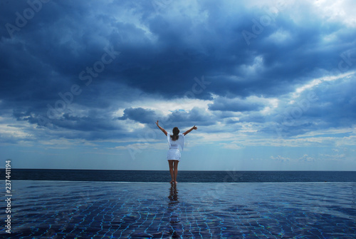 Girl in angel wings and white clothing standing on the edge of the pool in front of the sea with raised hands. Sky full of of clouds and dark blue ocean on the background. Pool in front. Photo © Julia Whitecrow