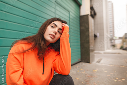 Street portrait of a stylish girl in an orange hoodie, sitting on the street and posing on the camera. Beautiful girl in street clothes sitting on the street.