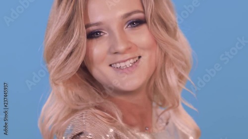 Close-up of a beautiful blonde in a golden dress. She winks with her eye, flirting and smiling. The background is blue.