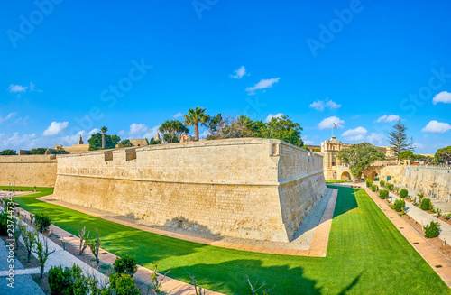 The huge St Peter's Bastion of Mdina fortress, surrounded by green lawn and trimmed bushes of local park, stretching along historic moat, Malta. photo