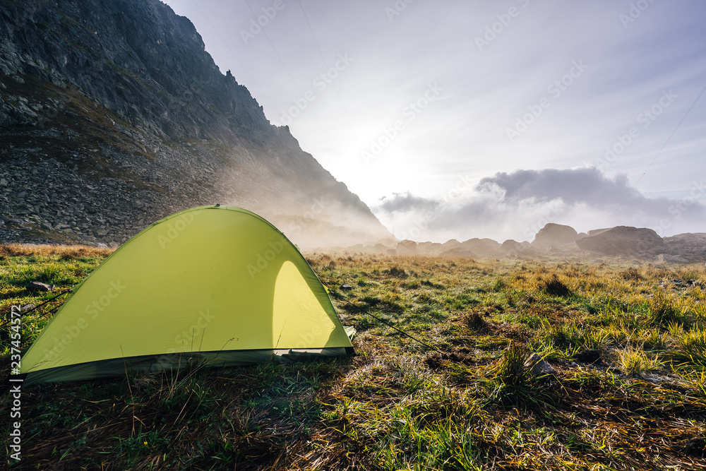 A green tent pitched in a green grass in the alpine like mountain landscape. Morning sunrise mist and colors, high peak behind. Camping outdoors.