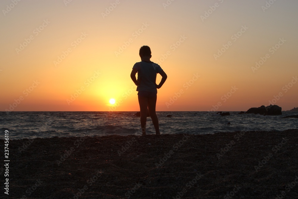 Silhouette of alone young boy with hands on waist staring at the sunset on the beach