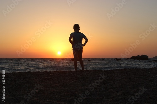 Silhouette of alone young boy with hands on waist staring at the sunset on the beach