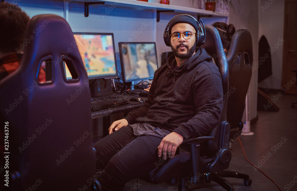 Fotografie African American bearded guy wearing hoodie and cap sitting on a gamer chair and looking at a camera in a gaming club or internet cafe
