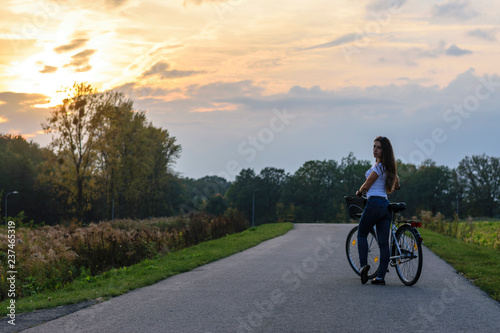 Young brunette girl with a bicycle in a park alley lit by sunset light.