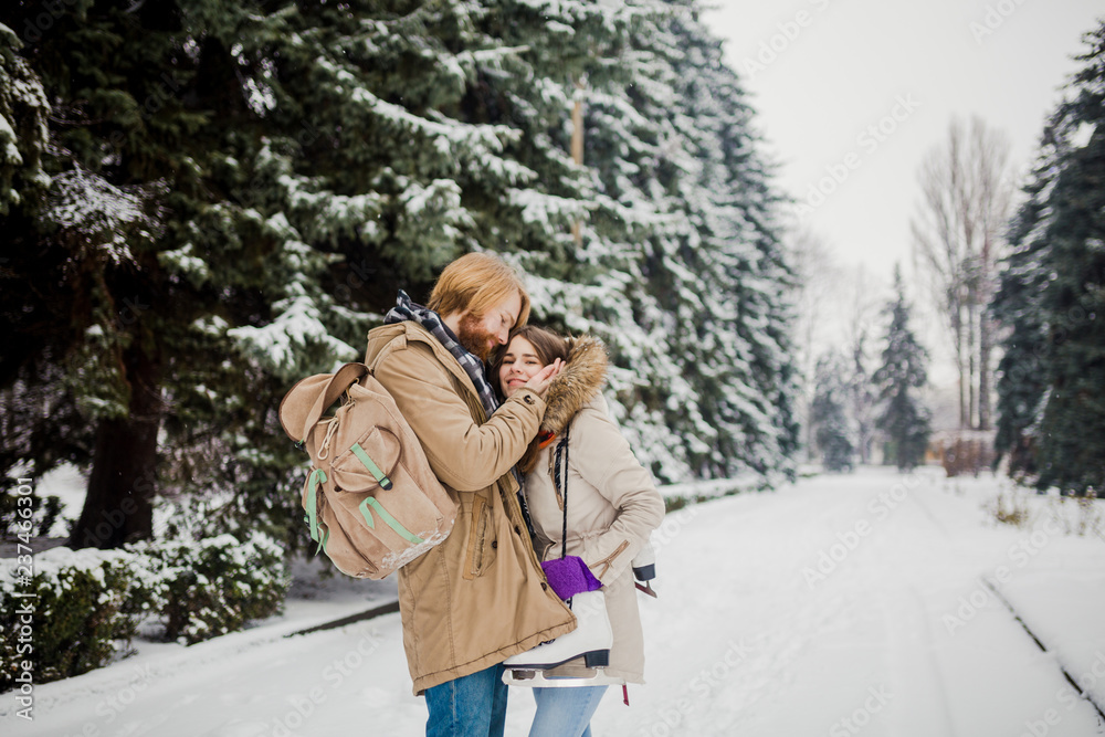 Date young couple in winter. Young Caucasian man with beard and long hair holding in hands the hands of girl with knitted gloves and warming and kissing them in snow park in winter is Valentines Day Date young couple in winter. Young Caucasian man with beard and long hair holding in hands the hands of girl with knitted gloves and warming and kissing them in snow park in winter is Valentines Day