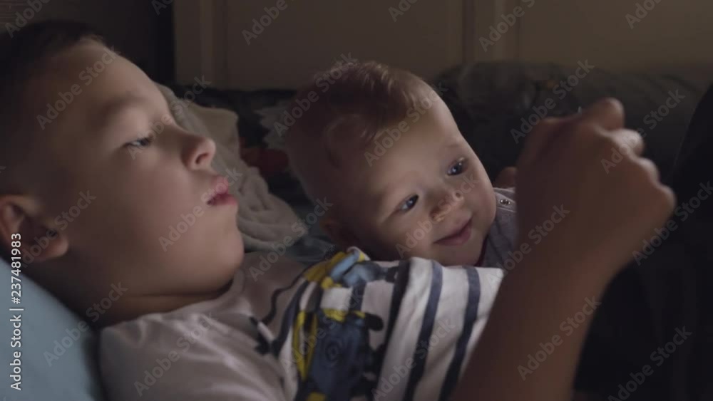 Medium shot of an older brother watching something on tablet with younger baby sister at home Medium shot of an older brother watching something on tablet with younger baby sister at home