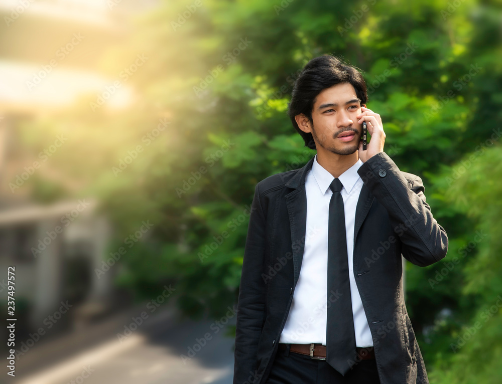Smiling young handsome bearded businessman walking and talking on smartphone over green city and office building background, business, technology and people concept. Copy space. Smiling young handsome bearded businessman walking and talking on smartphone over green city and office building background, business, technology and people concept. Copy space.