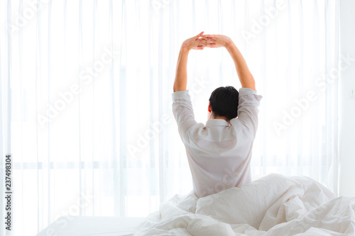 Black hair of young business asian man sitting near the big white window while stretching his arms on bed after waking up with sunrise at morning, back view.