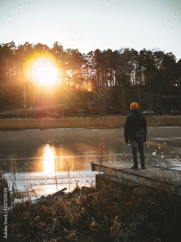 Blond man in winter jacket and yellow hat stands on the bridge on the pier and looks into the distance At the landscape at sunset, icy lake/ river, the sun sets in the woods behind the trees
