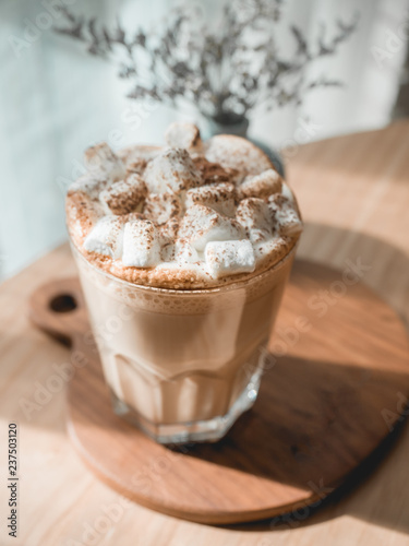 One glass of hot coffee with marshmallow on top serve on white tray on wood table next to bright window in morning.