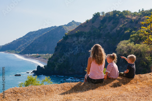 Family vacation lifestyle. Happy mother, kids on hill with scenic view of high cliffs, fishers village on black beach. Children looking at blue sea. Bukit Asah is popular travel destination in Bali.