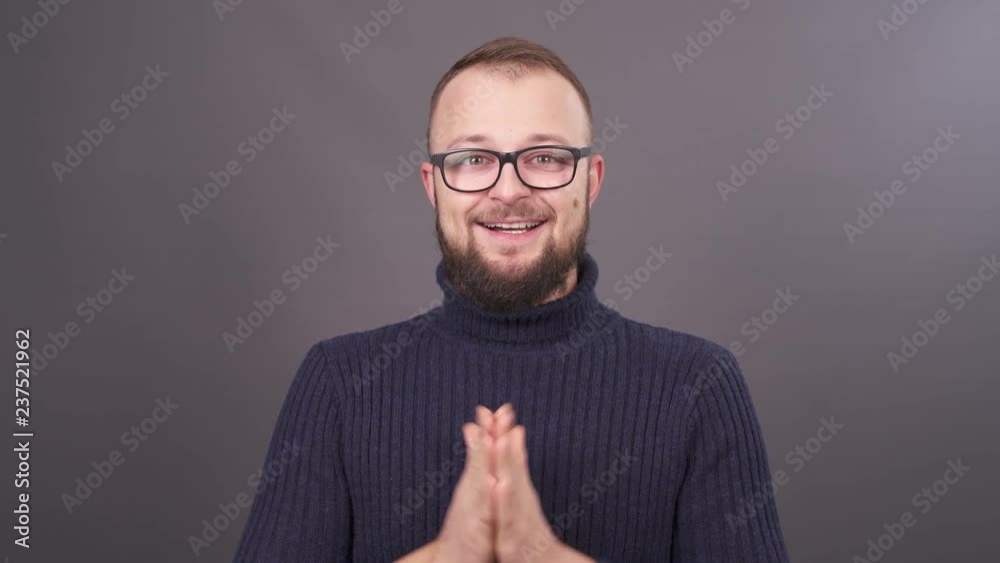 Close-up portrait of a bearded man in glasses rubs ones hands. Isolated on grey background.