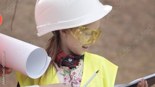 Two children girls in construction helmets looking at white sheet of paper or drawing and smiling