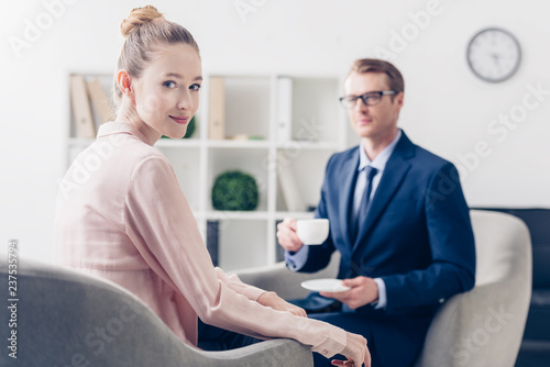 handsome businessman holding cup of tea, beautiful journalist looking at camera in office