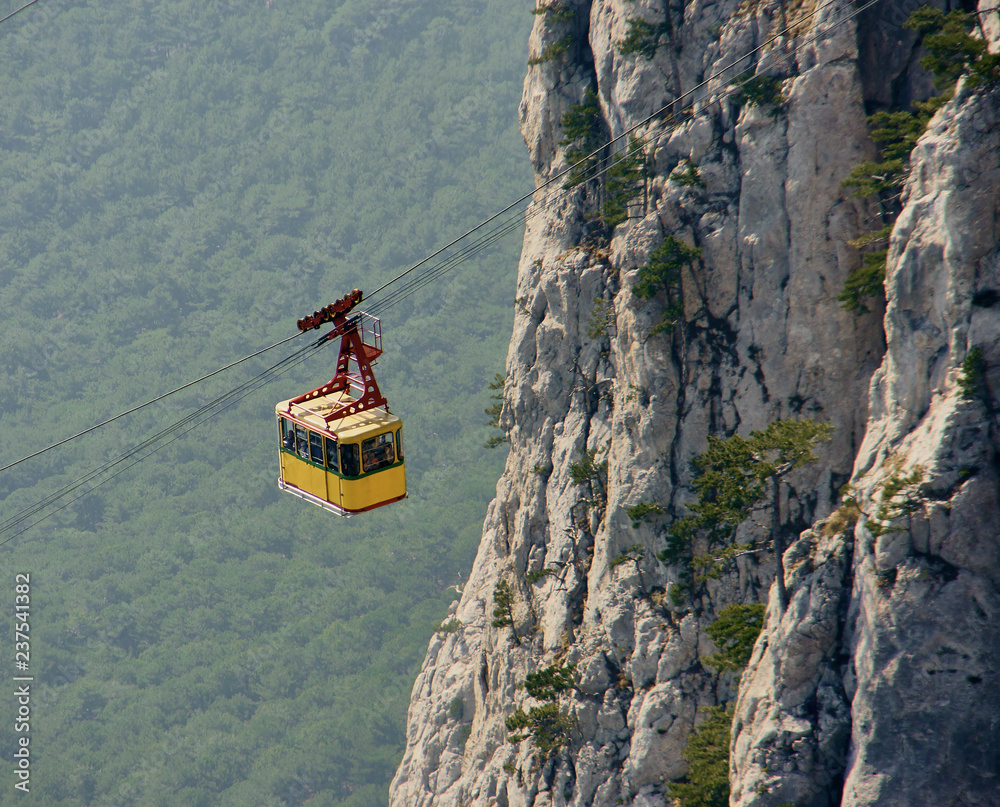 View of the cable car on the highest mountain of Crimea (Mount Ai-Petri). Cabin with passengers climbs on the rock. Evergreen pines grow on a cliff against a blurred background of trees.