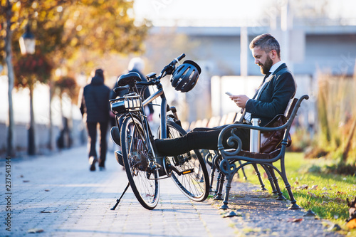 Businessman commuter with bicycle sitting on bench in city, using smartphone. © Halfpoint