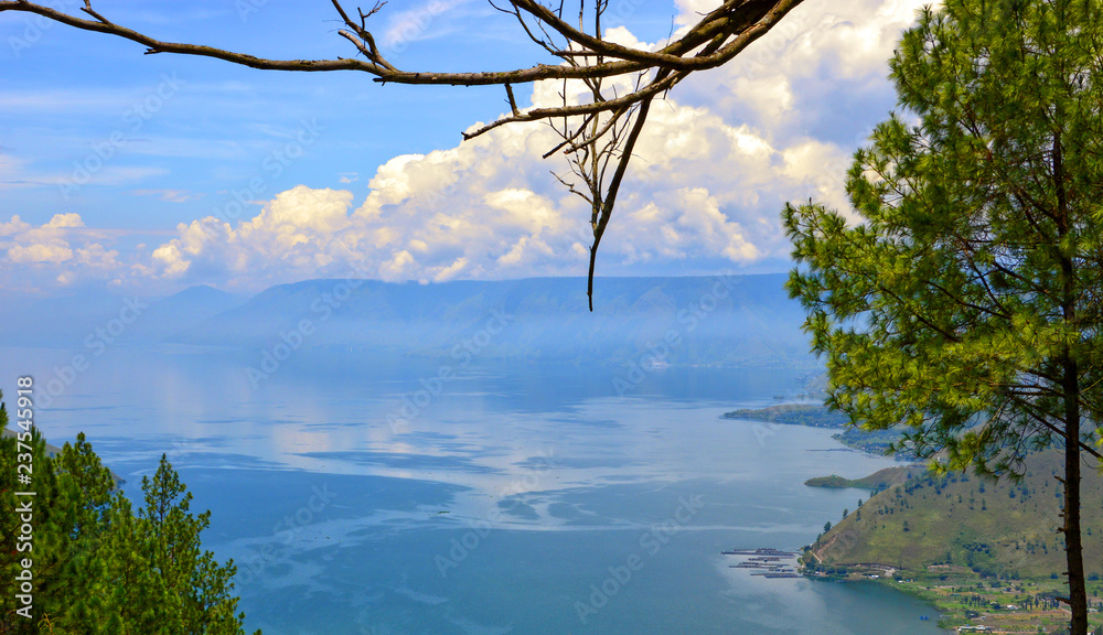 Fototapeta premium Stunning view of the Lake Toba, Indonesia. Tree branch and pine trees in the foreground. Lake Toba or Danau Toba is a huge crater lake in Sumatra.