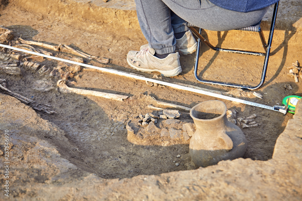 Real archaeological excavation. Archaeologist draws the tomb with human bones, part of skeleton, skull and ceramic jar in the ground. Outdoors Real archaeological excavation. Archaeologist draws the tomb with human bones, part of skeleton, skull and ceramic jar in the ground. Outdoors