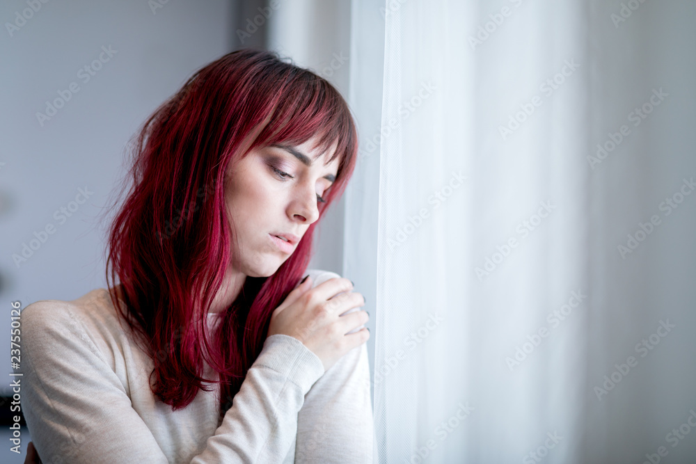 Depressed young woman near window at home thinking about unpleasant experiences Depressed young woman near window at home thinking about unpleasant experiences