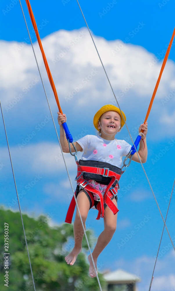 A little girl jumps high on a trampoline with rubber ropes against the blue sky and white clouds. Adventures and extreme sports. Concept of summer vacation, jumping.