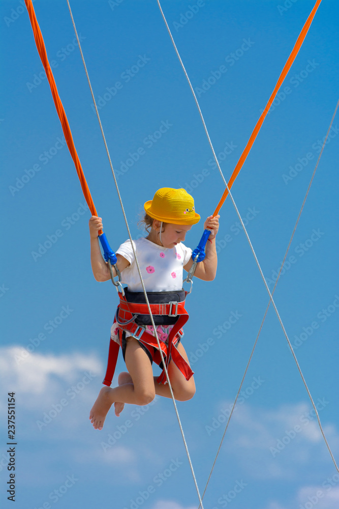 A little girl jumps high on a trampoline with rubber ropes against the blue sky and white clouds. Adventures and extreme sports. Concept of summer vacation, jumping.