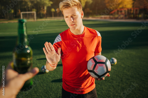 Fototapeta Naklejka Na Ścianę i Meble -  Picture of oyung player showing stop sign to human that offers him bottle of alcohol. Guy hold ball in hand. He stand on lawn.