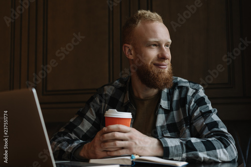 Happy bearded hipster man freelancer with emotional smiling face using laptop computer, working, planning project, thinking, drinking coffee in modern cafe. Remote job. Successful business 