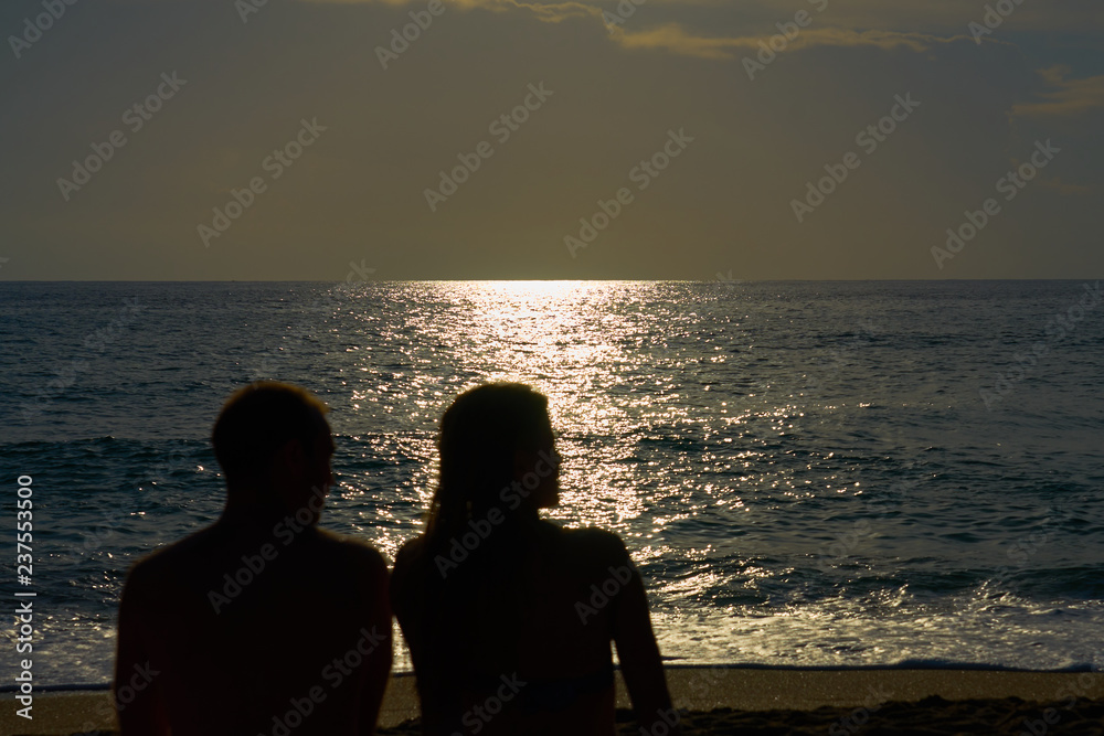 Silhouettes of a couple, man and woman are sitting on a sandy beach and looking at the sunset on the sea on a vacation. Copy space.    