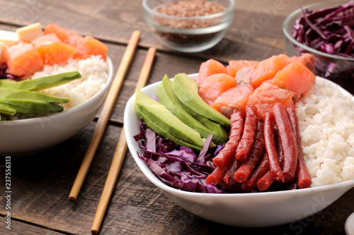 poke bowl Hawaiian food. a plate of rice, salmon, avocado, cabbage and cheese, next to sesame and soy sauce. on a brown wooden table.