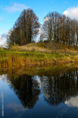 Autumn landscape with colorful trees, yellow grass and river. Reflection in river