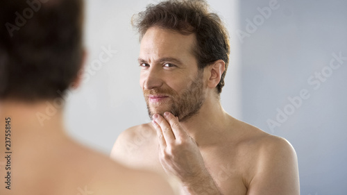 Topless male critically looking at his beard in mirror, morning ritual, bathroom