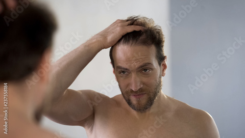 Worried man looking in mirror at his bald patches, hair loss problem, hygiene