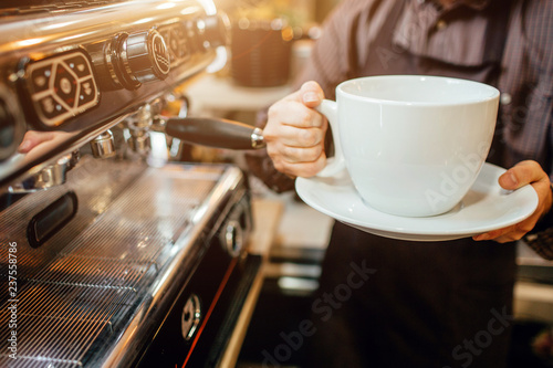 Cut view of barista holding huge white cup of coffee in hands. He stands in kitchen at coffee machine.