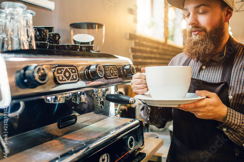 Calm and peaceful young man stand at coffee machine in kitchen and smells coffee from huge white cup. He looks at it with pleasure. photo