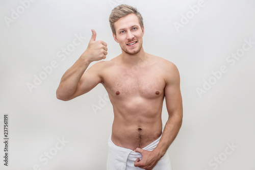 Positive young man looking on camera. He show big thumb up and hold towel in hand arounf hips. Guy is happy. Isolated on white background. photo