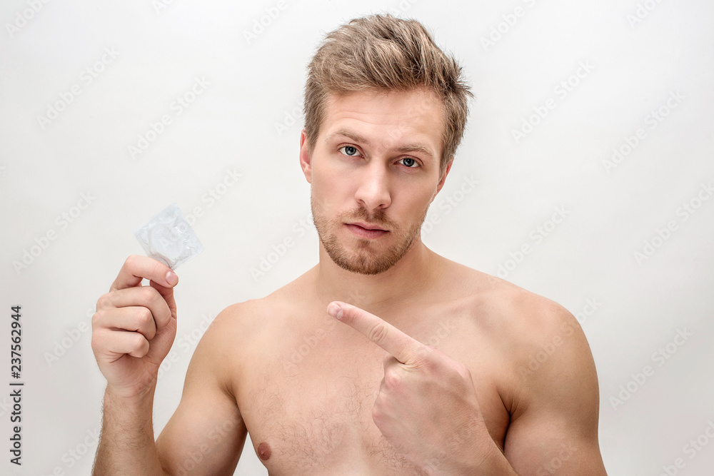 Young man hold condom in hand. He point on it. Guy look straight. Isolated on white background. Young man hold condom in hand. He point on it. Guy look straight. Isolated on white background.