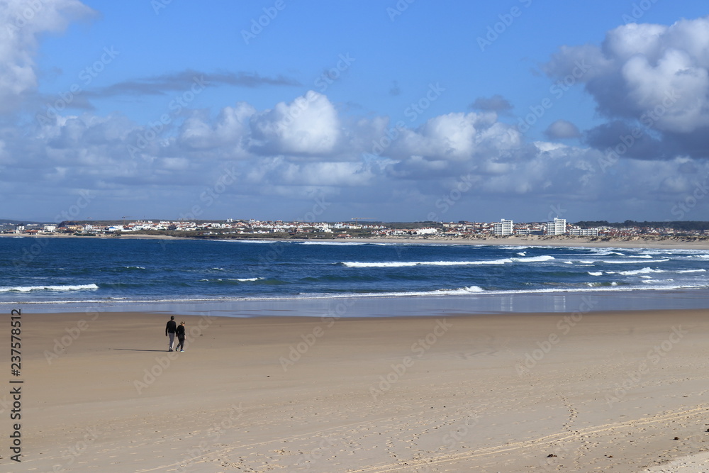 ouple walking on beach sand with dark blue sea and cloudy sky in background, Peniche