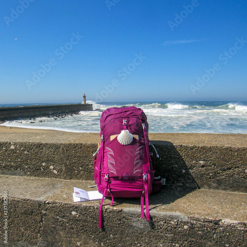 Pink backpack with scallop shell symbol of pilgrim at concrete wall with ocean with huge white waves and blue sky on background photo