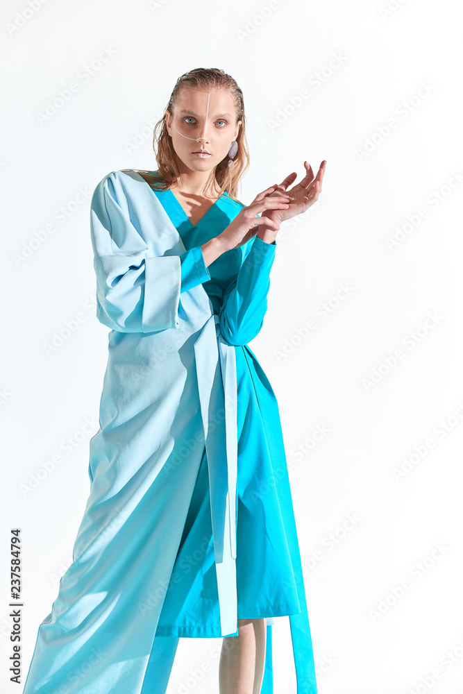 Fashion portrait of young blonde woman with blue eyes and painted white lines on face, dressed in turquoise dress. White background
