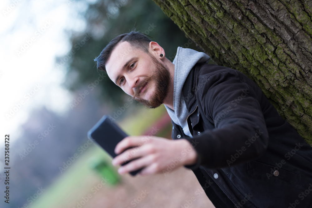 Fototapeta premium A handsome young hipster man leaning against a tree and taking a selfie in the park.