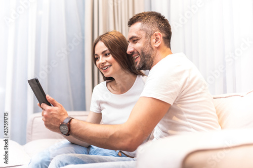 Profile of hilarious twosome sitting on couch and looking at device screen with wide smile