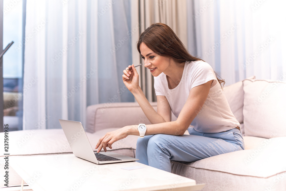 Happy female person is sitting on sofa and typing on laptop. She looking at screen with wide smile. Copy space on left side