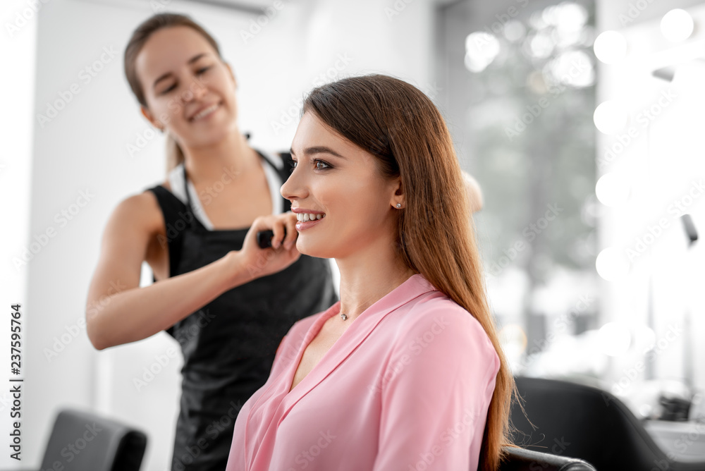 Waist up photo of beautiful smiling woman is sitting in beauty salon while female stylist making hair styling Waist up photo of beautiful smiling woman is sitting in beauty salon while female stylist making hair styling