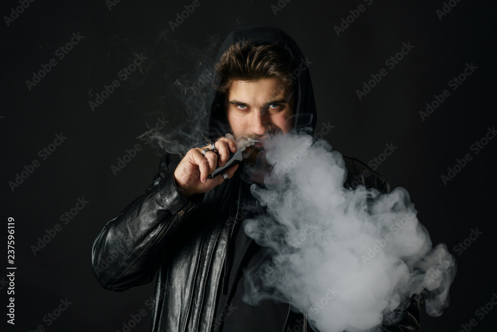 Man with vaping mod exhaling steam at black studio background. Bearded guy smoking e-cigarette to quit tobacco. Vapor and alternative nicotine free smoking concept Man with vaping mod exhaling steam at black studio background. Bearded guy smoking e-cigarette to quit tobacco. Vapor and alternative nicotine free smoking concept