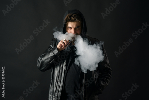 Man with vaping mod exhaling steam at black studio background. Bearded guy smoking e-cigarette to quit tobacco. Vapor and alternative nicotine free smoking concept