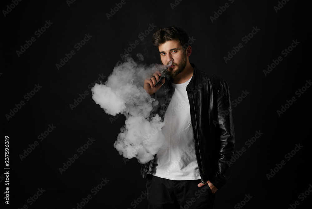 Man with vaping mod exhaling steam at black studio background. Bearded guy smoking e-cigarette to quit tobacco. Vapor and alternative nicotine free smoking concept Man with vaping mod exhaling steam at black studio background. Bearded guy smoking e-cigarette to quit tobacco. Vapor and alternative nicotine free smoking concept