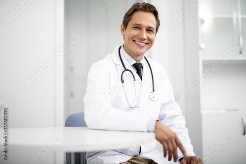 Emotional confident practitioner in white coat smiling and putting one hand on the table while sitting and looking