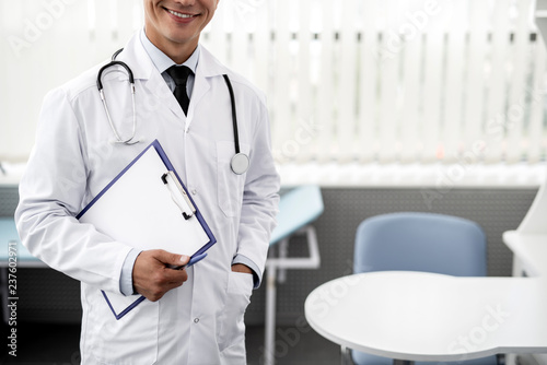 Cheerful emotional doctor wearing white medical clothes and smiling while holding clipboard and having stethoscope on his neck