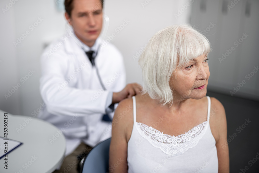 Fototapeta premium Calm aged lady sitting on the chair and turning her head while general practitioner listening to her with his stethoscope